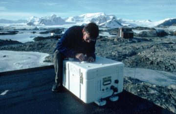 SAOZ - observes spectra of UV and visible sunlight scattered from the zenith sky at twilight, to measure total column ozone and Nitrogen dioxide. SAOZ (Systeme d'Analyse d'Observations zenithales) is in a weather-proof box and looks up through the simple quartz window in its lid. Derek Oldham is seen here installing SAOZ on the roof of the ozone loft in March 1990. Faraday station was occupied by BAS from 1947 to 1996, it was transferred to the Ukraine in February 1996 and renamed Vernadsky. SAOZ was moved to Rothera when Faraday was transferred to the Ukraines