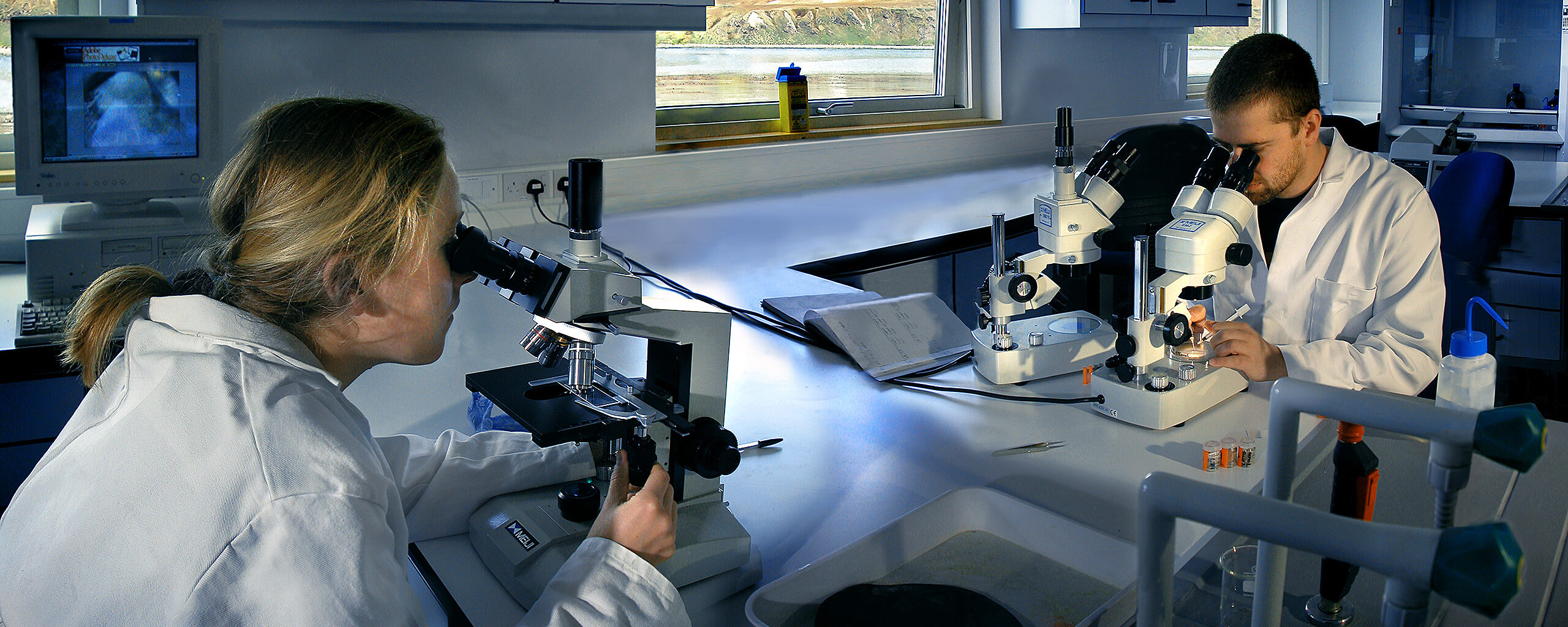 Two scientists using microscopes. King Edward Point is visible out of their laboratory window