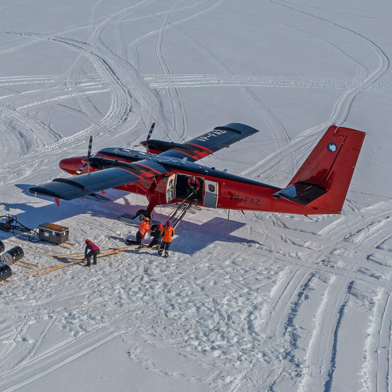 A red Twin Otter aircraft loading cargo including fuel drums at Gromits Creek, Antarctica.